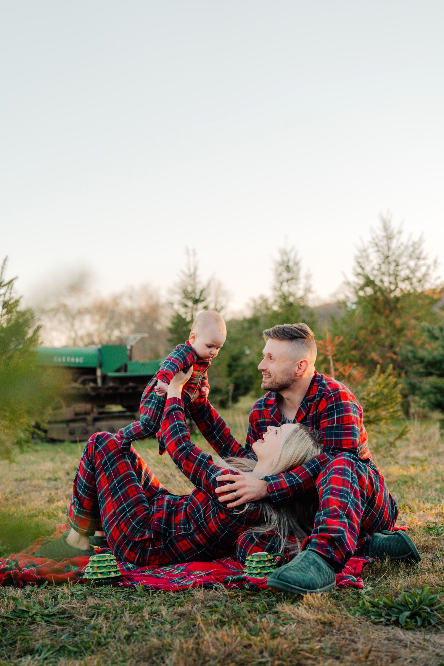 Family wearing matching red plaid pajamas relaxing on picnic blanket outdoors during holiday photoshoot.