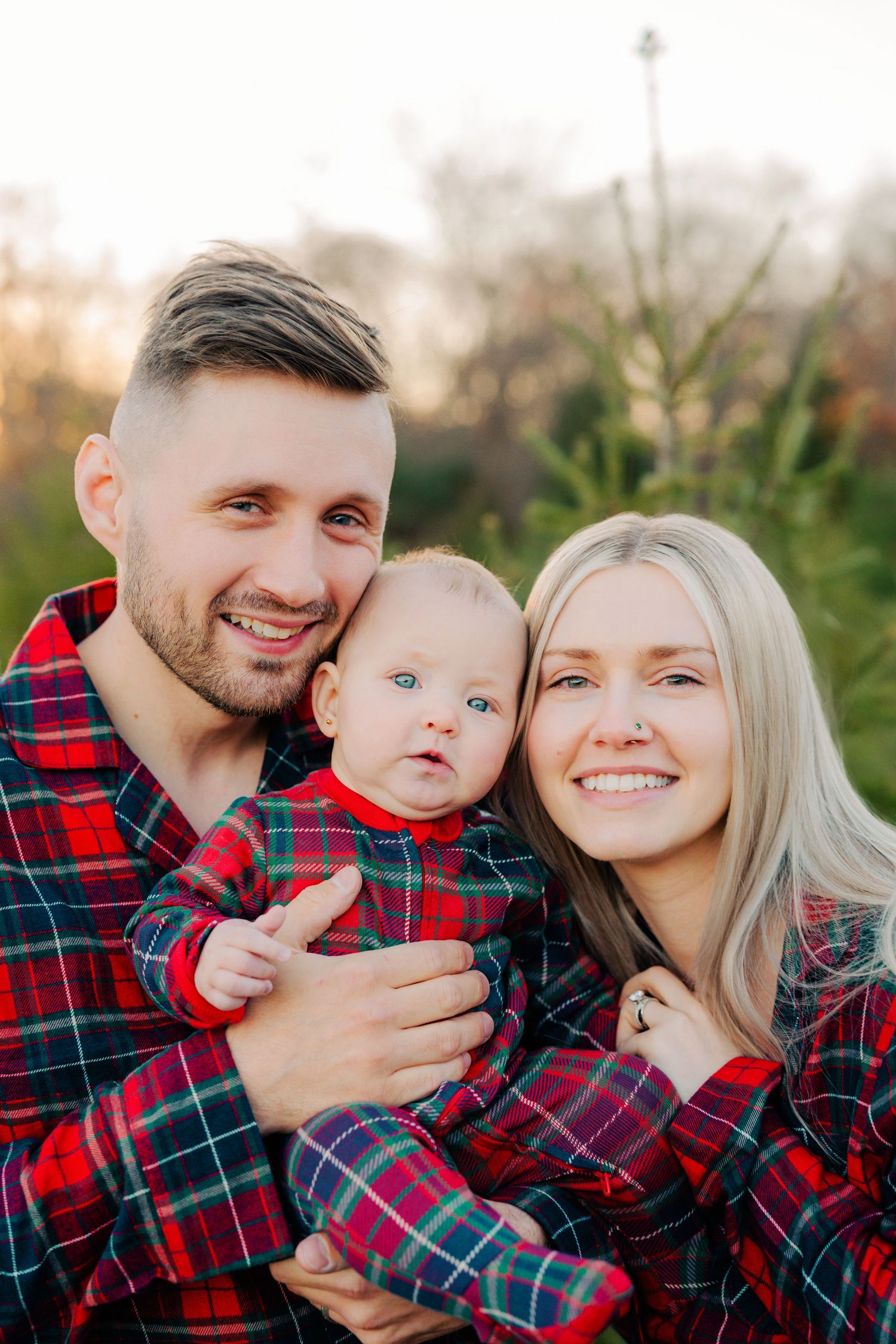 A family wearing matching red plaid flannel shirts poses together outdoors during an autumn photo session.