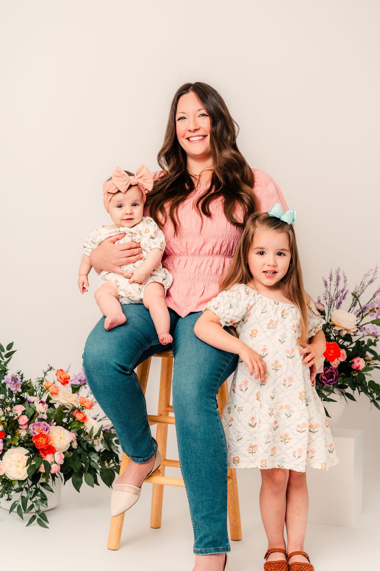 A woman in a pink top and jeans sits on a stool holding a baby, with a young girl in a floral dress beside her.