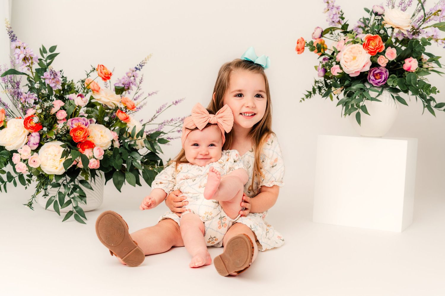 Two young sisters in floral dresses pose together surrounded by colorful flower arrangements on a white background.