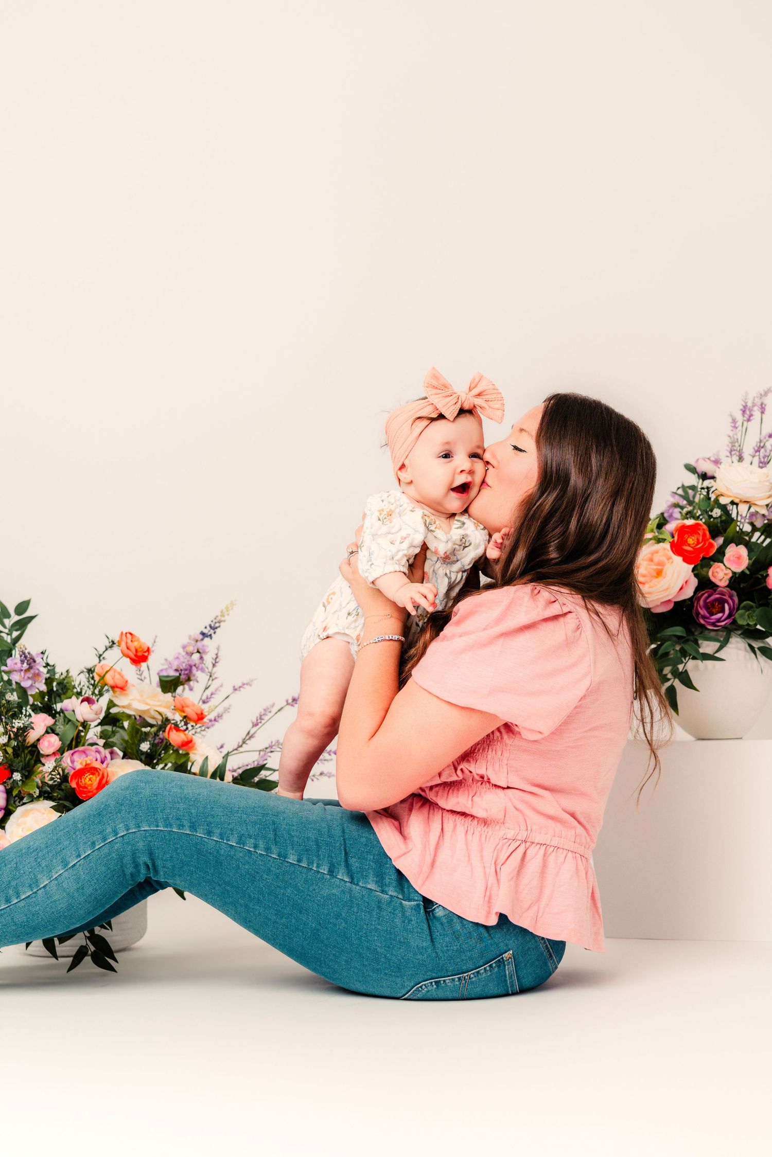 A mother in a pink top and jeans sits on the floor kissing her baby girl surrounded by colorful flower arrangements.