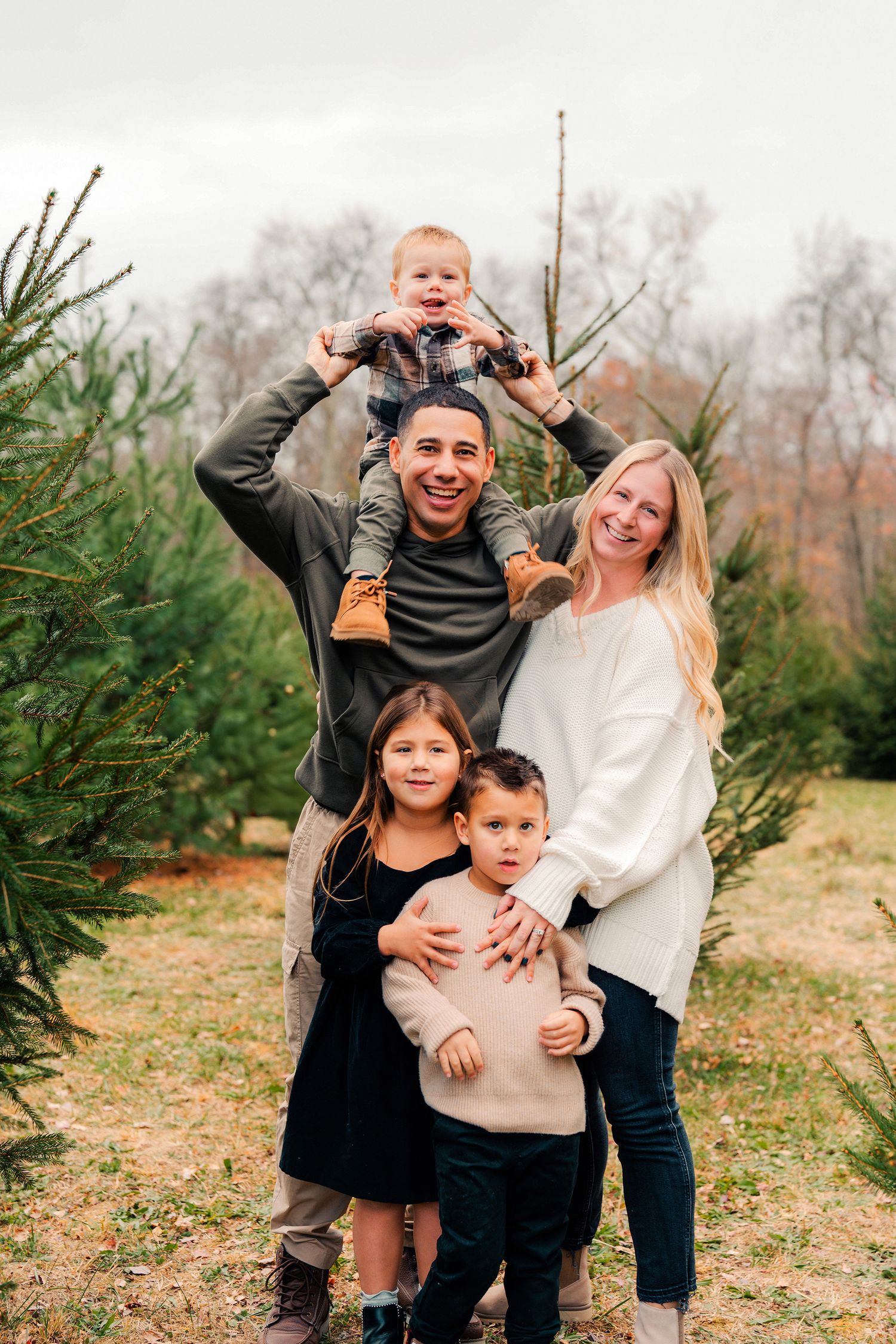 Happy family posing together at a Christmas tree farm with evergreen trees in the background.