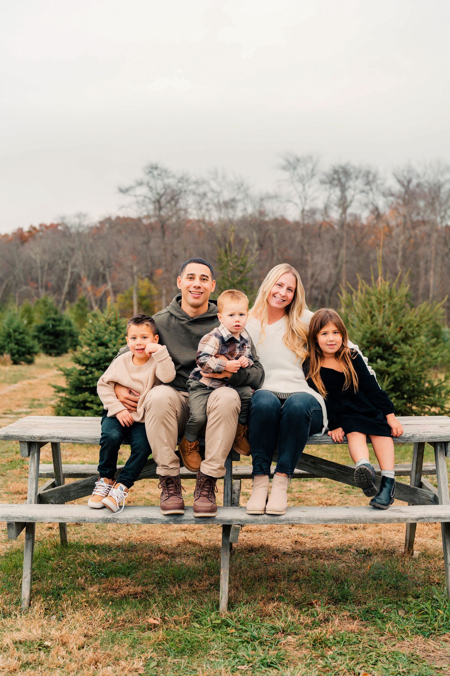 A family sits together on wooden picnic bench steps at a Christmas tree farm surrounded by evergreens on an autumn day.