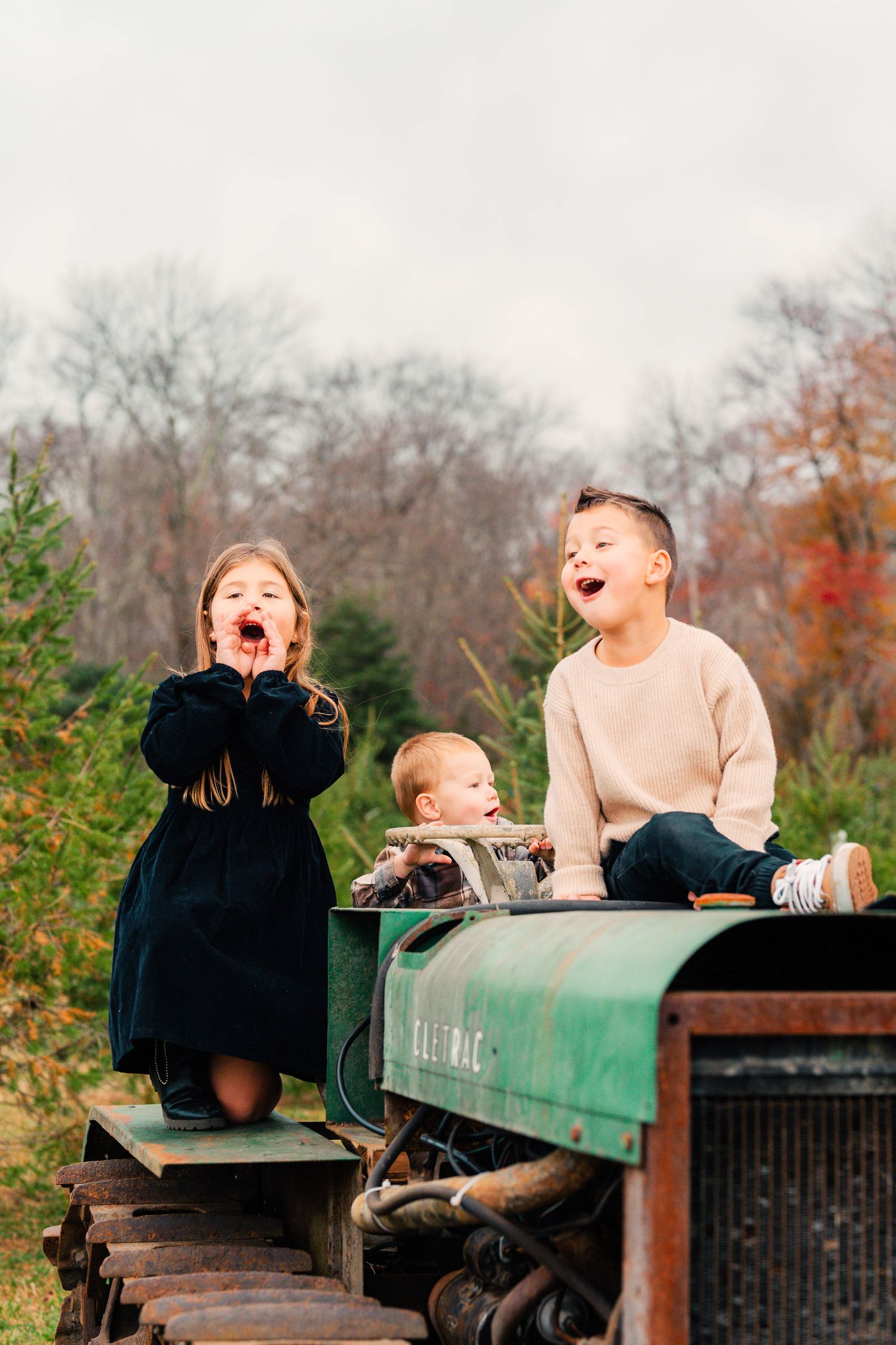 Three children sitting and laughing together on a vintage green tractor during autumn with fall foliage in background.