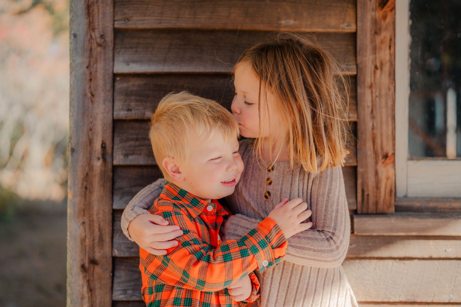 A tender moment captured between a child in an orange plaid shirt and a companion in a beige sweater by a rustic wooden structure.