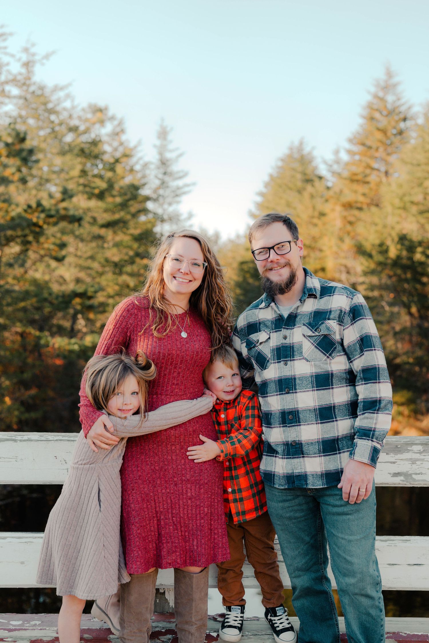 A family poses together on a wooden deck surrounded by evergreen trees during a sunny autumn day.