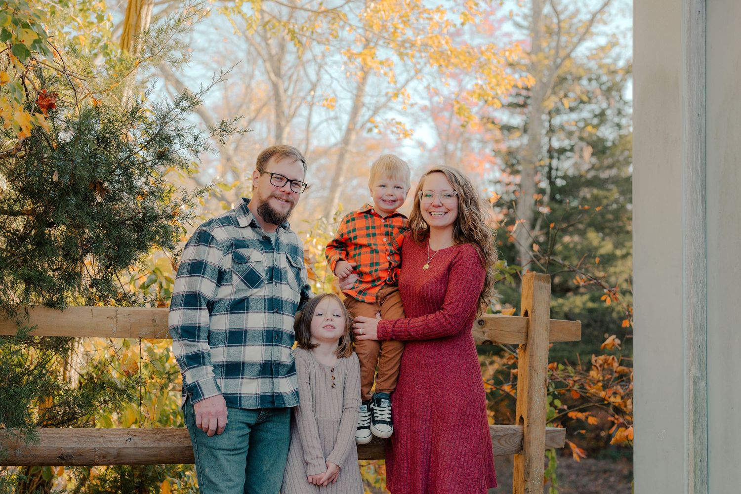 A family in autumn attire poses together on a wooden deck surrounded by fall foliage with warm golden light.