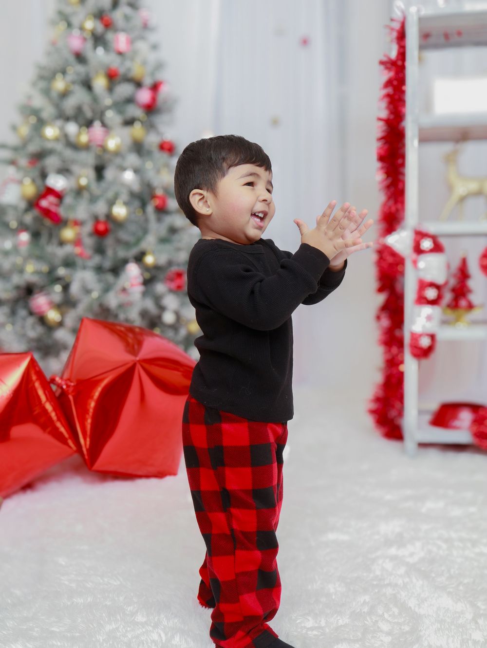 A child in black sweater and buffalo plaid pants claps excitedly by a Christmas tree.