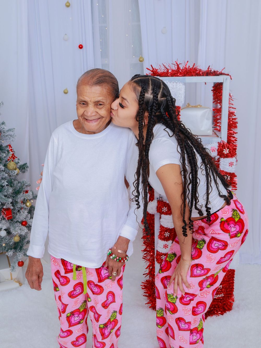 Two people in matching pink heart pajamas pose together next to a decorated Christmas tree and red tinsel.