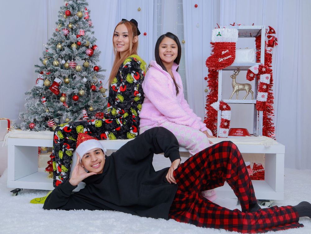 Three people in holiday outfits pose playfully on a white bench near a decorated Christmas tree.