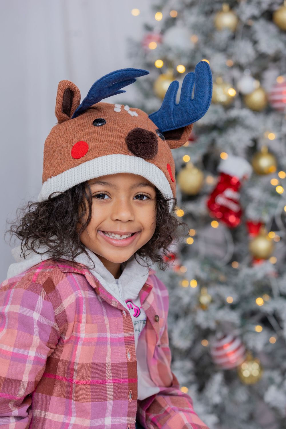 A child wearing a reindeer winter hat smiles in front of a decorated Christmas tree with twinkling lights.