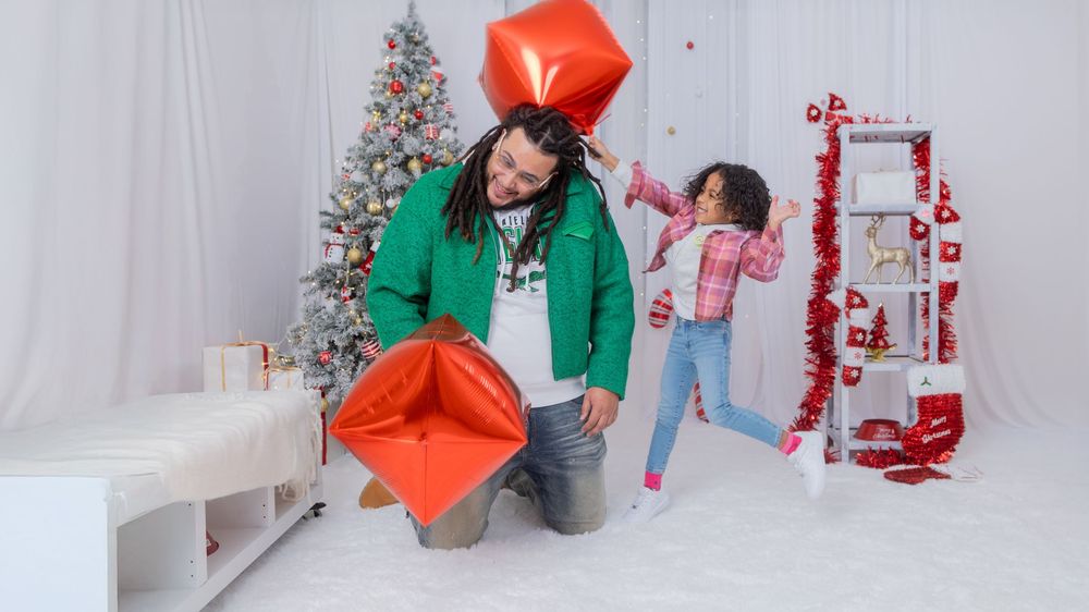 A playful holiday photoshoot scene with a large red gift box prop and decorated Christmas tree.