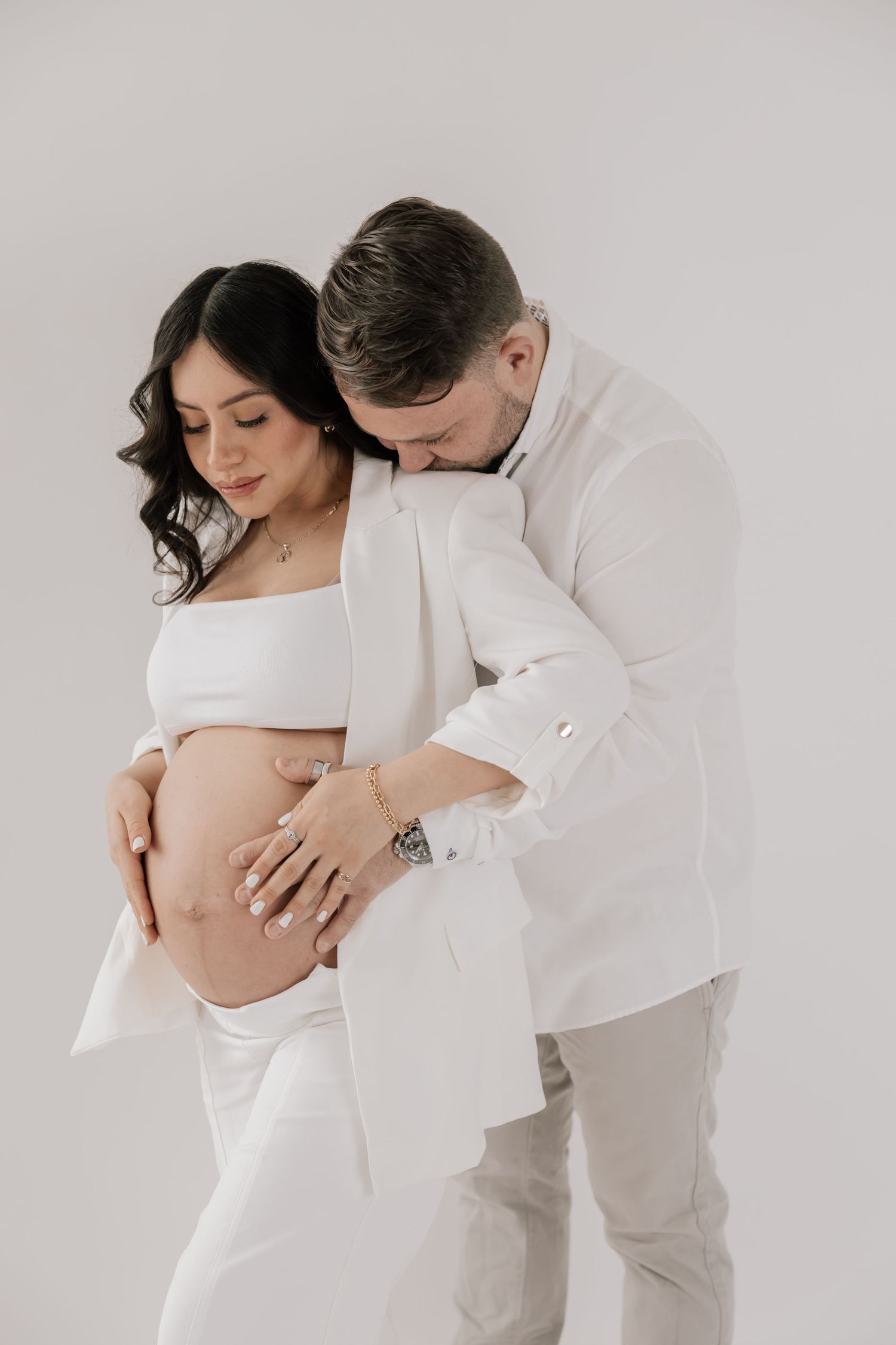 Intimate maternity photo of a couple in white outfits embracing against a light background.