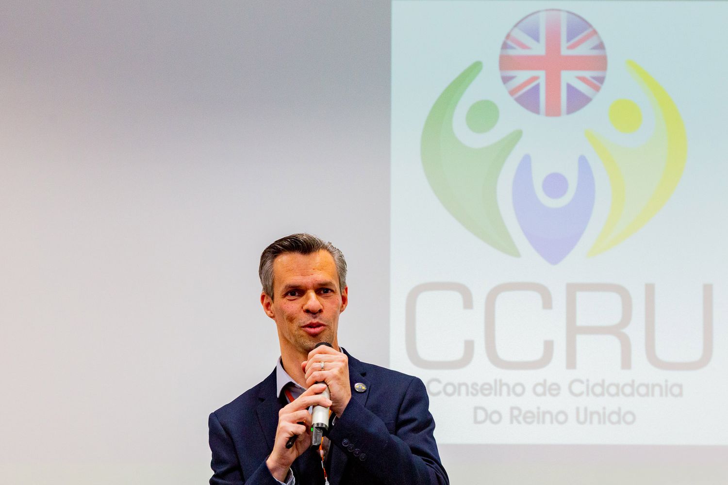 A man in a blue suit holds a microphone while speaking at a podium with the CCRU logo visible in the background.