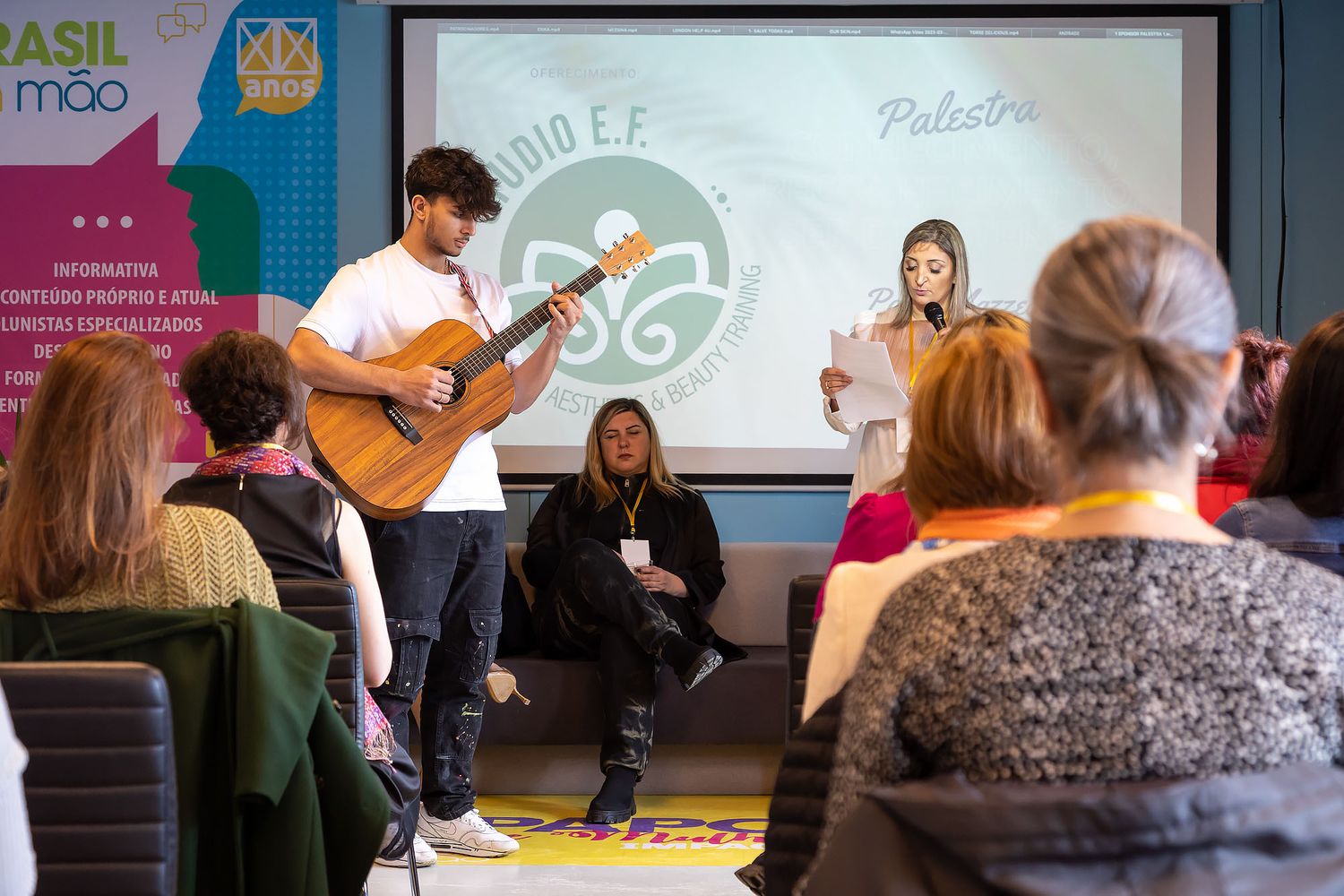 Musicians perform at an intimate venue with an acoustic guitar while audience members watch from chairs in a classroom setting.