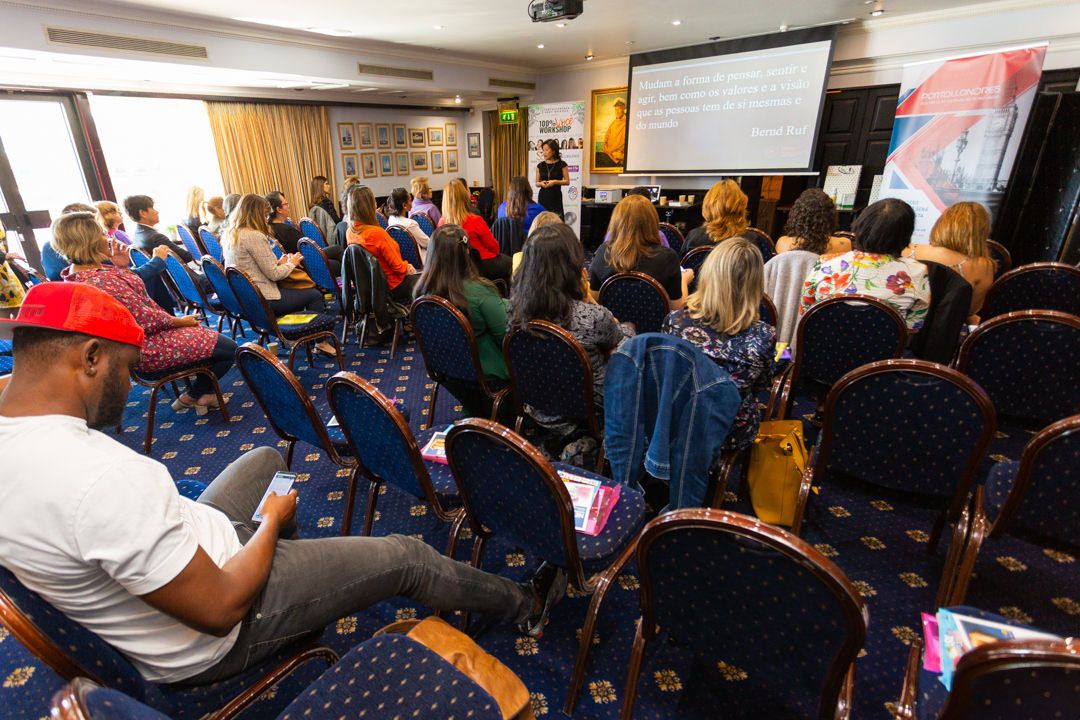 A crowded conference room filled with attendees sitting in rows of blue chairs facing a projection screen at the front.