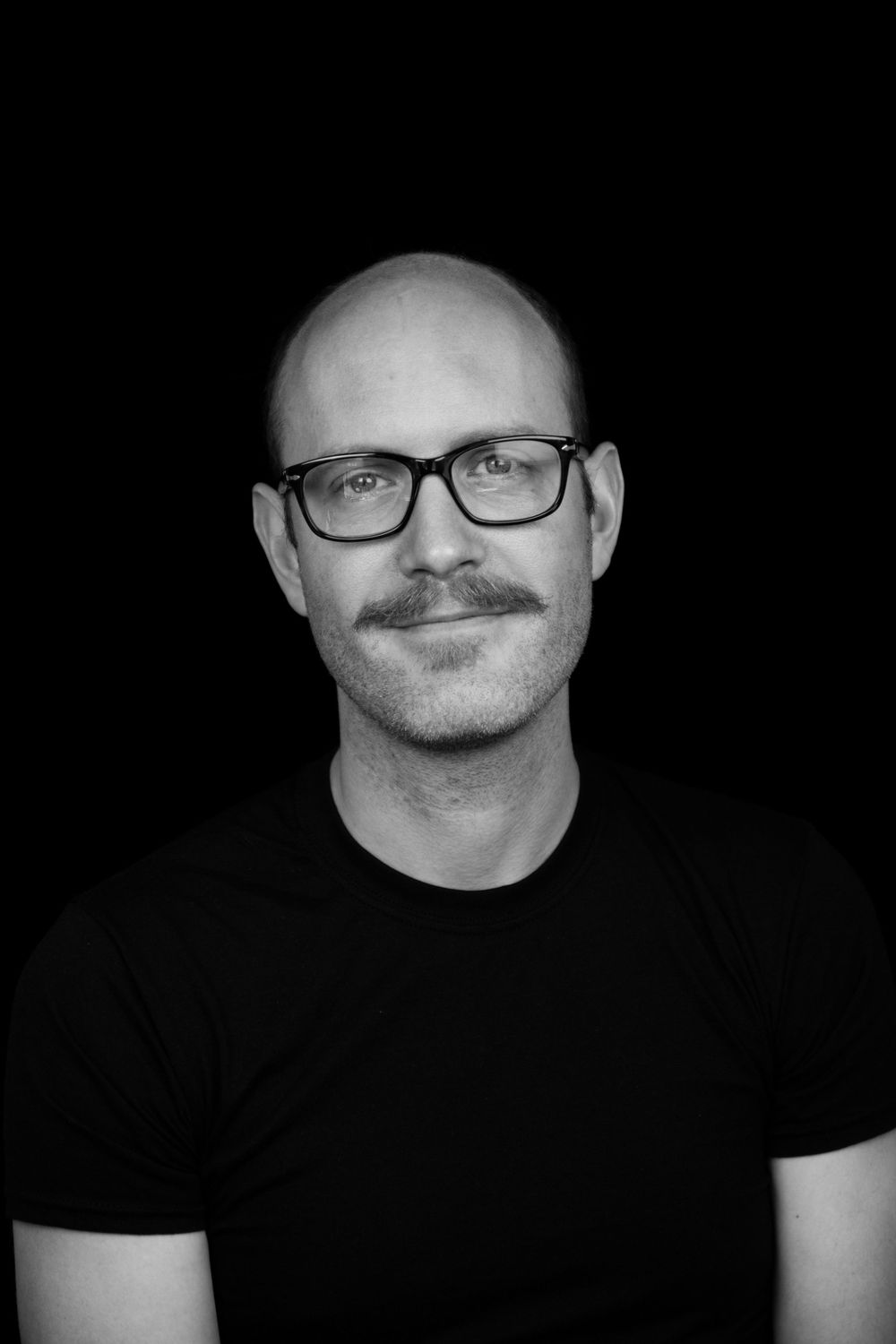 Black and white portrait of man in glasses and black shirt against dark background.