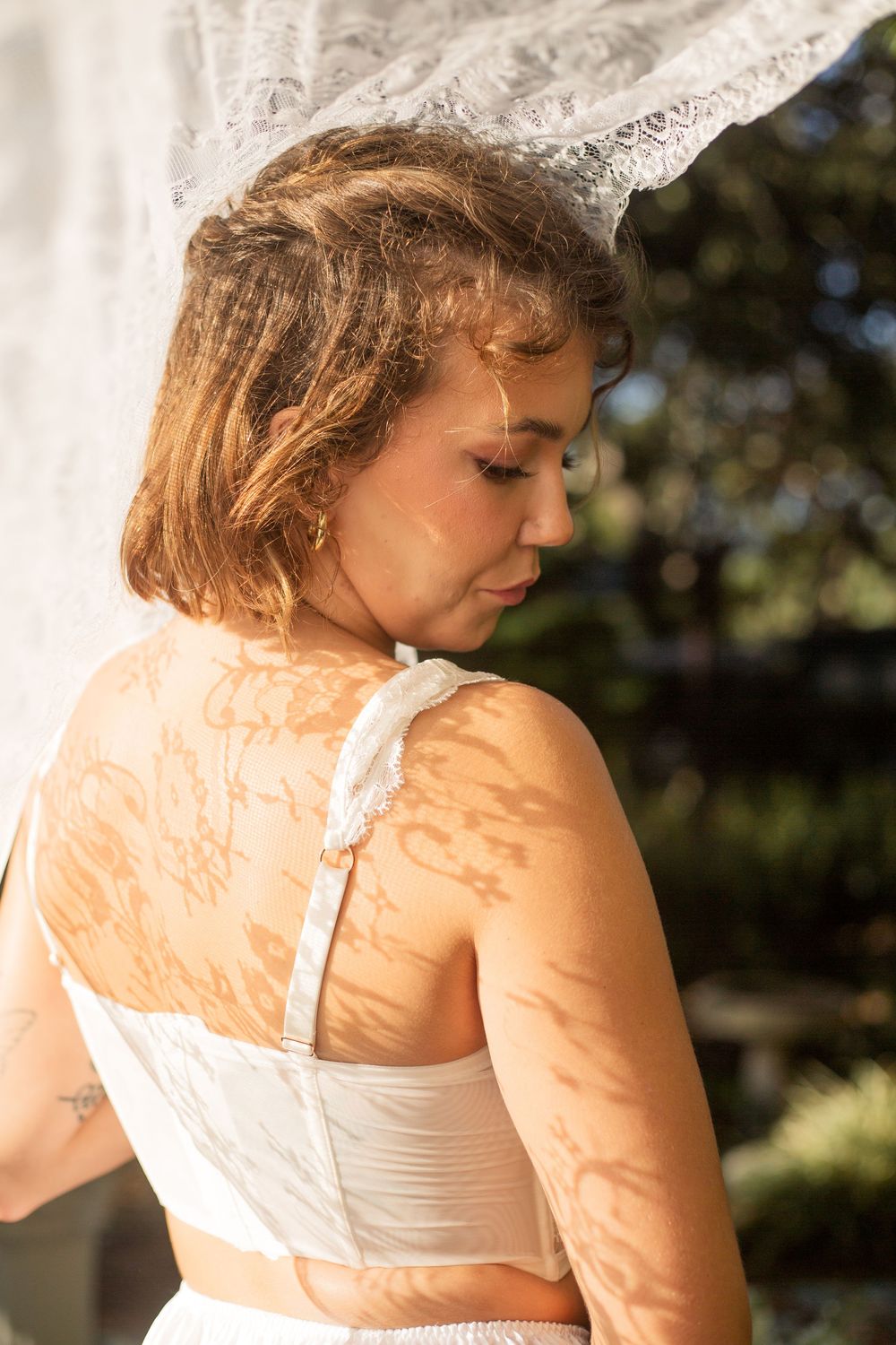A bride in a white dress stands in soft golden sunlight casting dappled shadows across her shoulders and back.