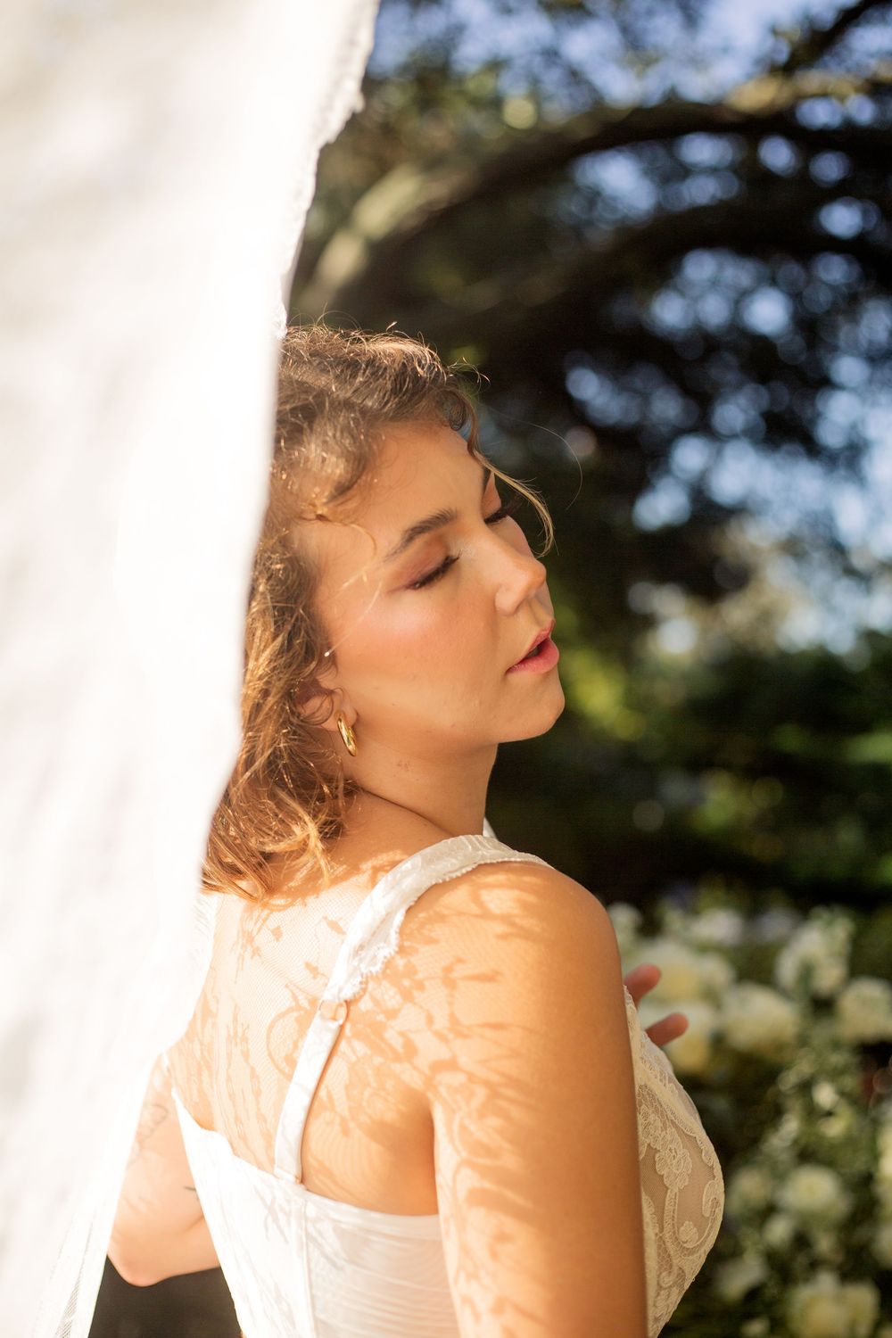 A woman in a white lace dress stands in golden sunlight with a thoughtful expression and closed eyes.