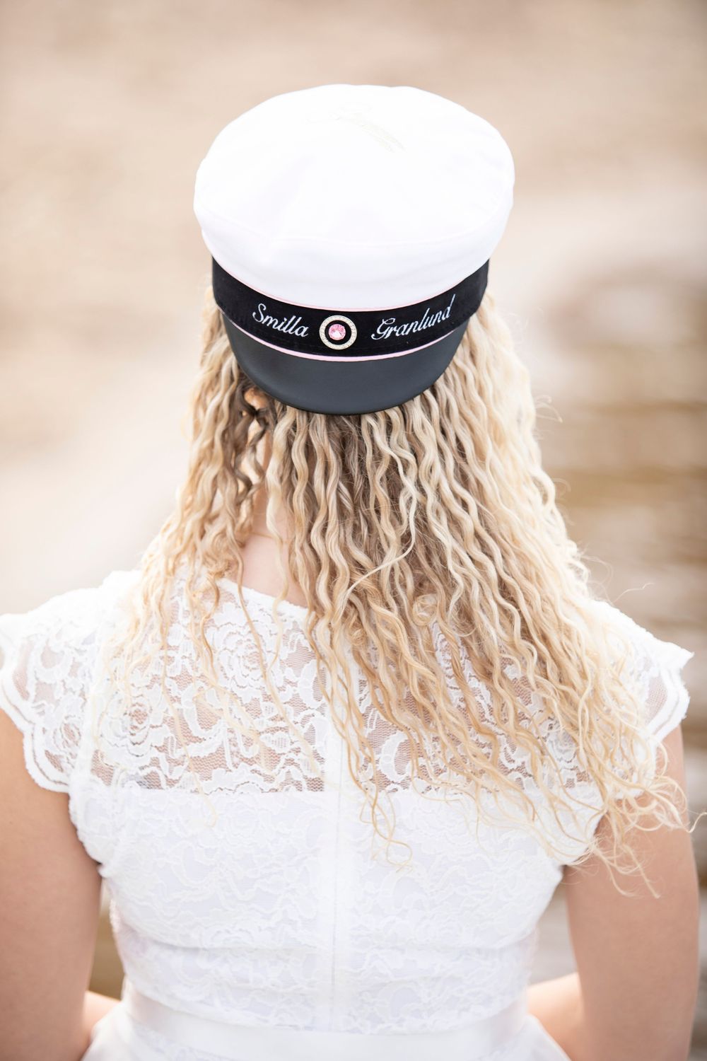 Back view of someone wearing a white lace dress and sailor cap with blonde curly hair falling down their back.