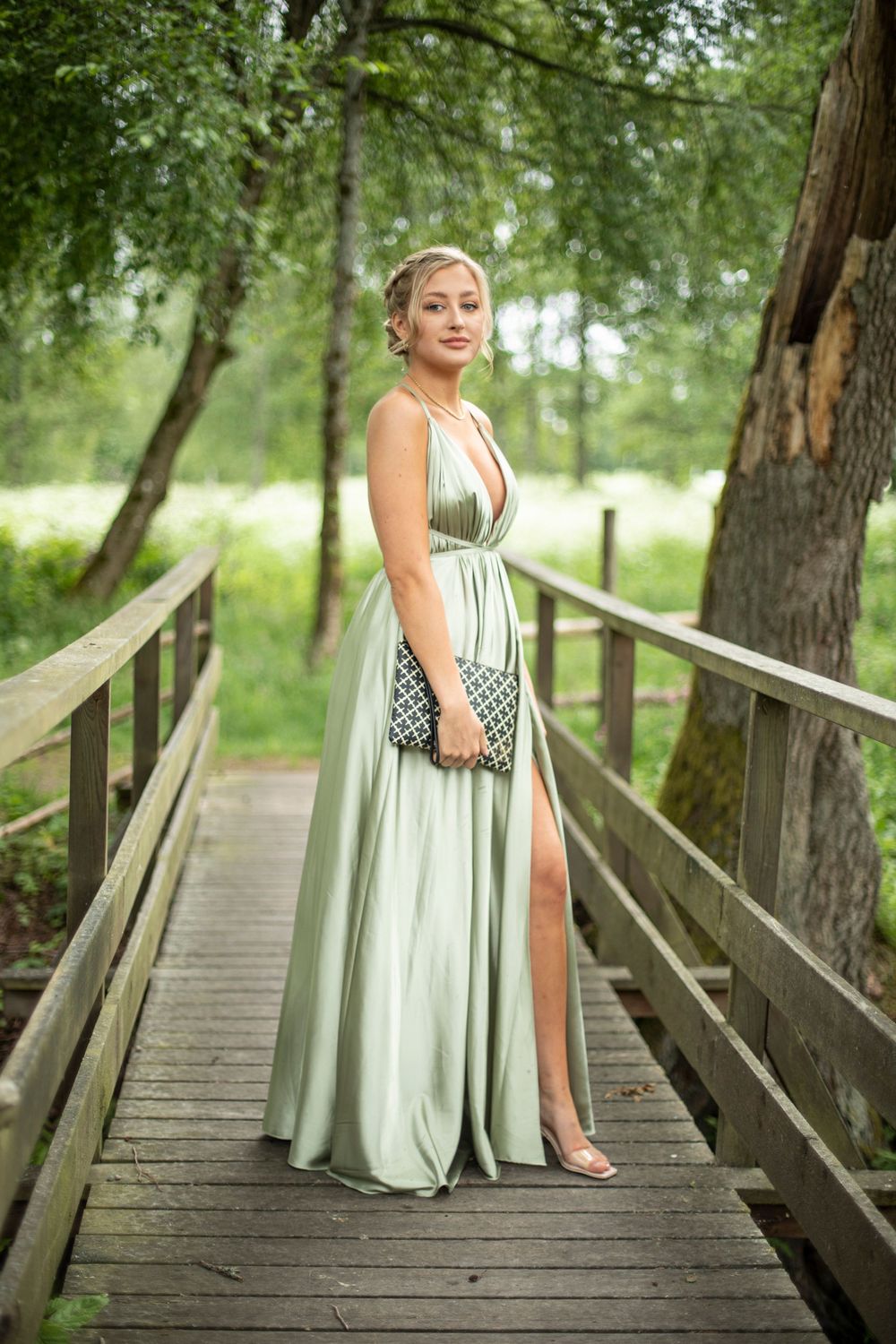 Woman in flowing mint green formal dress with slit poses on wooden bridge walkway surrounded by lush greenery.