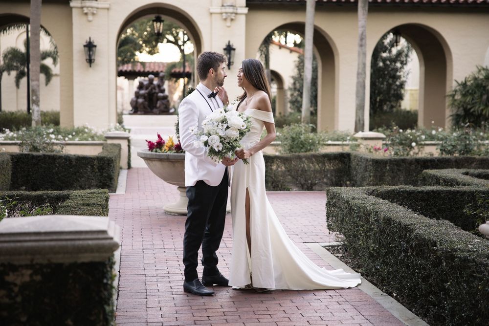 A couple in wedding attire share a romantic moment in a garden courtyard with arched architecture and manicured hedges.