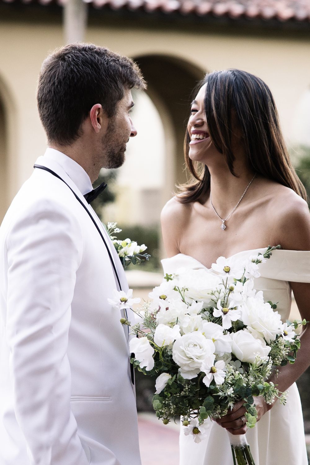 Wedding couple shares a joyful moment as bride holds stunning white bouquet while wearing an off-shoulder gown.
