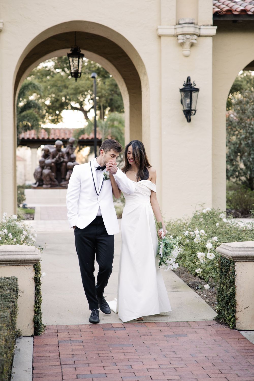 A couple in elegant wedding attire walks together under a Mediterranean-style archway with brick pavers and lanterns.