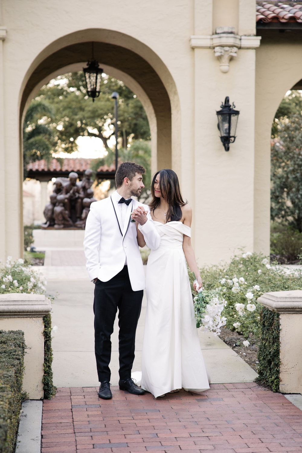 Couple in wedding attire standing together under an elegant archway at a Mediterranean-style venue with brick walkway.