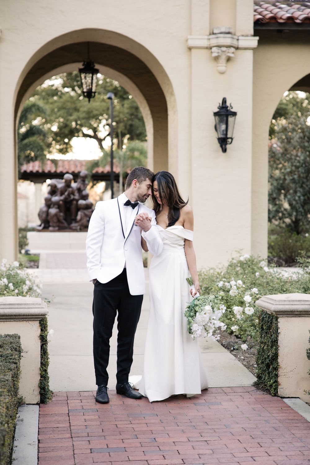 A couple in wedding attire shares an intimate moment under a Mediterranean-style archway with hanging lanterns and red brick pavers.