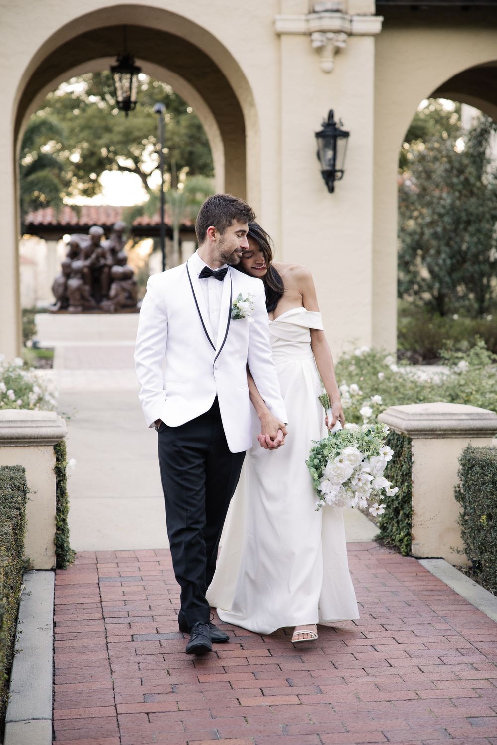 Elegant bride and groom share intimate moment while walking under archway at romantic outdoor wedding venue.