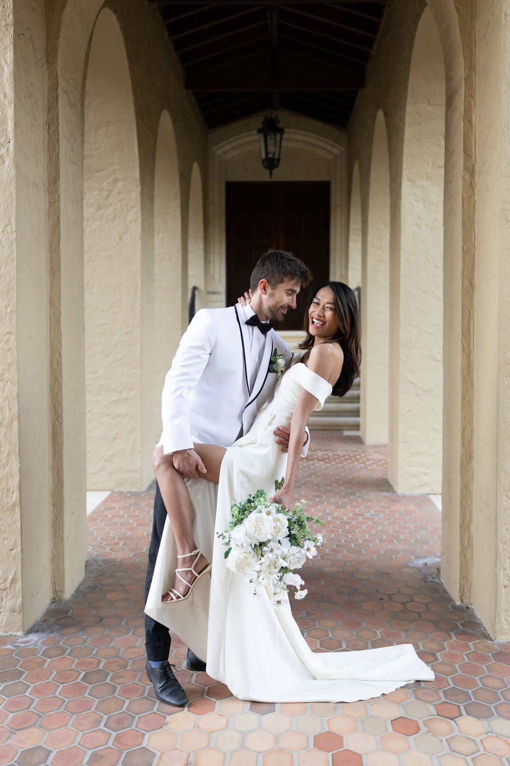 A romantic wedding photo of a couple in formal attire standing in an elegant arched hallway with a bridal bouquet.