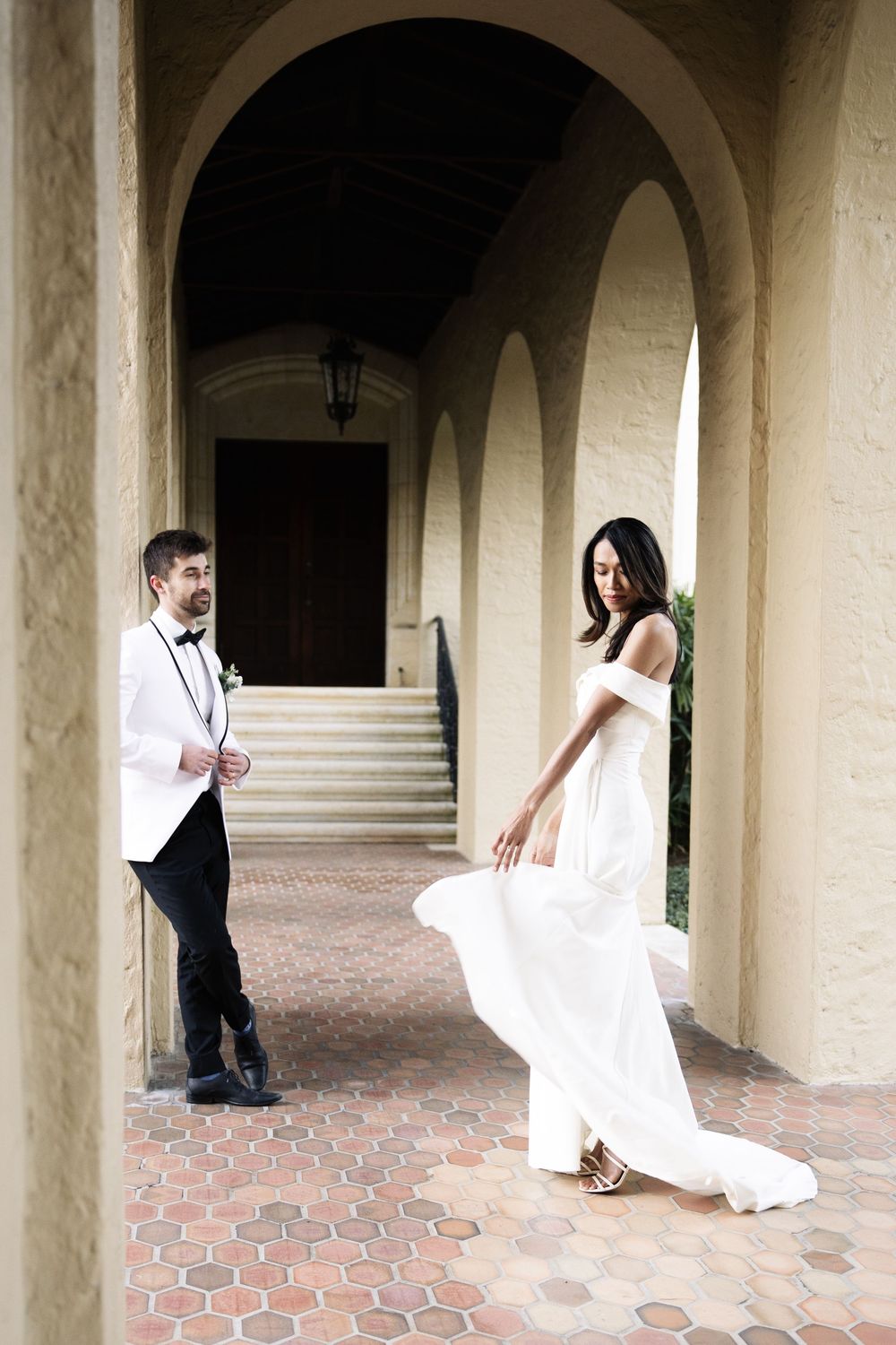 A couple in wedding attire pose playfully under arched columns on a brick paved walkway.