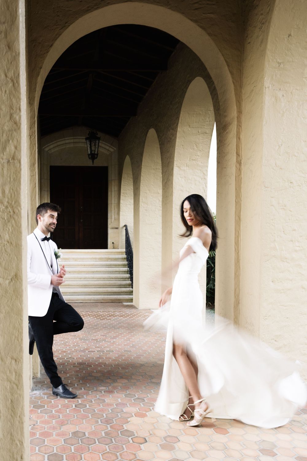 Couple in formal attire poses under elegant arched hallway with terra cotta flooring and soft natural lighting.