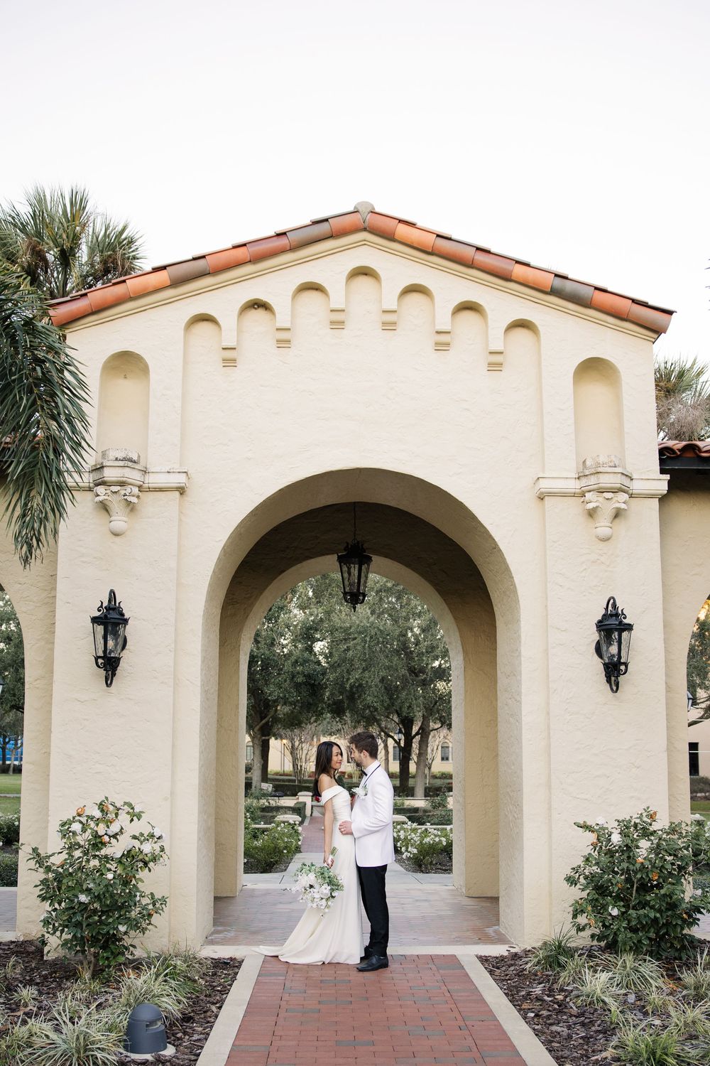 A couple stands under an ornate Mediterranean-style archway with lanterns and terracotta roof tiles in an elegant courtyard.