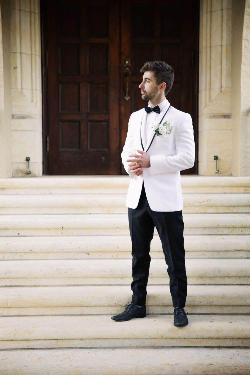 A groom in a white dinner jacket and black bow tie stands on stone steps outside an elegant wooden door.