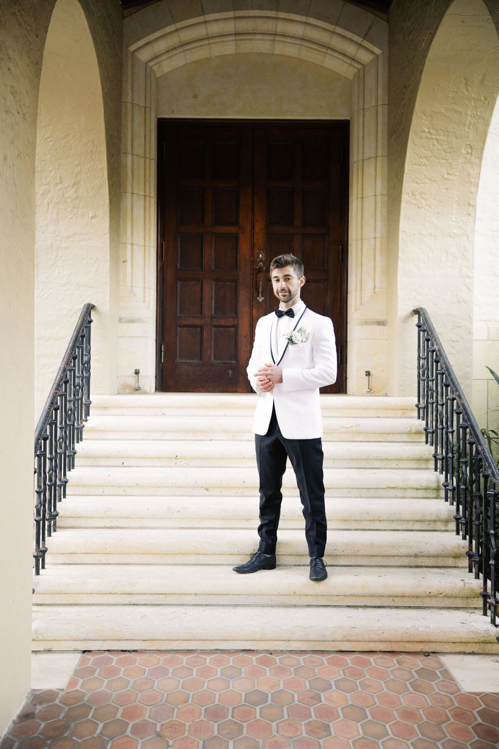 Someone in a formal white dinner jacket and black pants stands on stone steps with ornate iron railings in front of a wooden door.