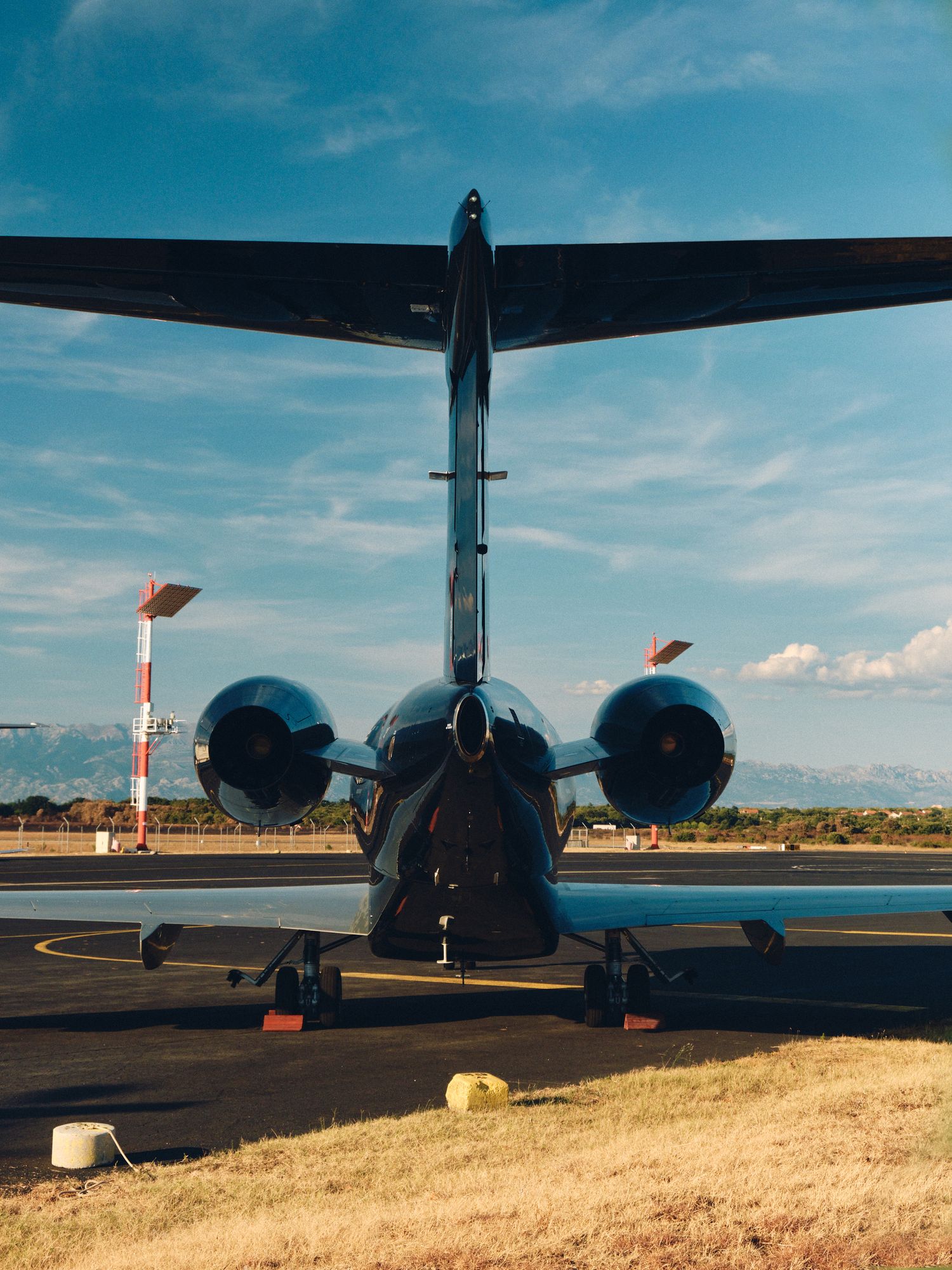 Rear view of a military transport aircraft with twin engines on tarmac against blue sky with windsocks visible.