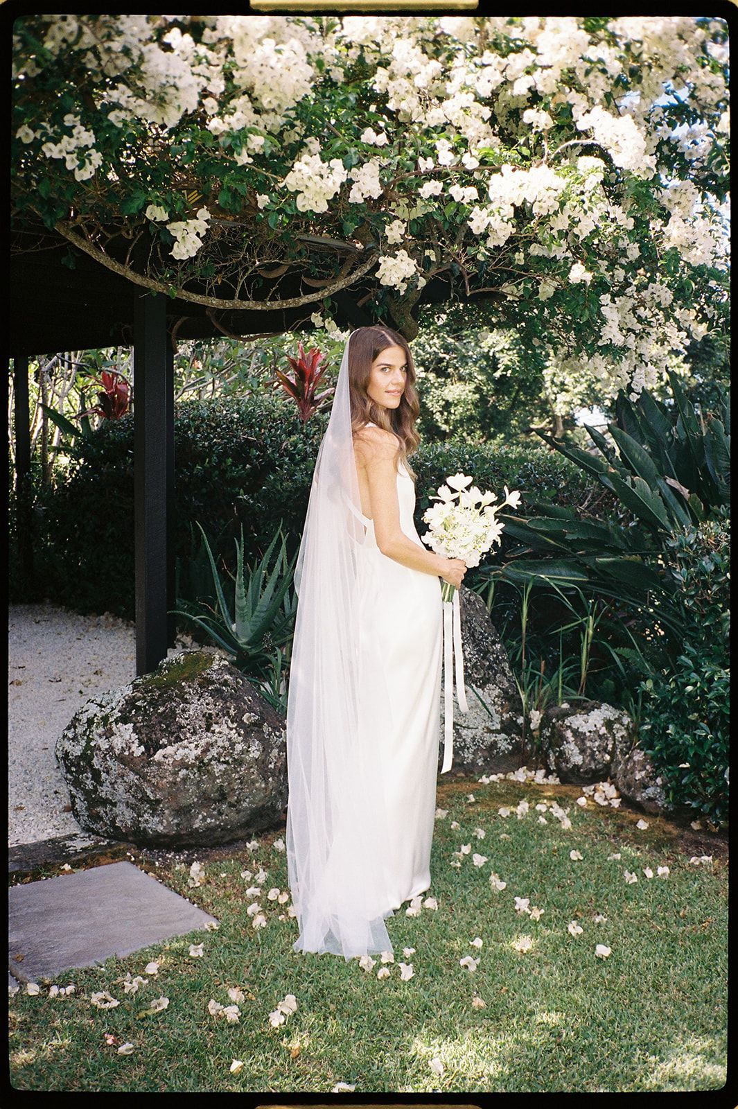 Bride in white gown and veil holds bouquet under flowering tree in lush garden with petals on ground.