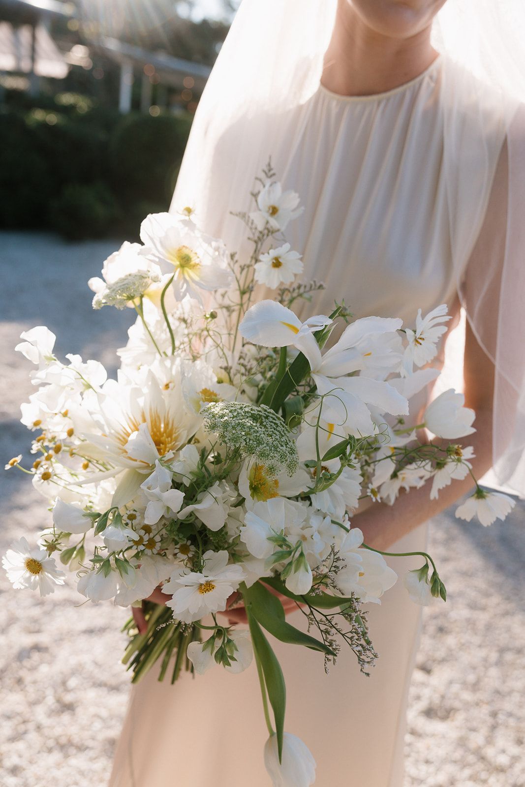 A delicate white wildflower wedding bouquet with cosmos and textural greenery held against a flowing white dress.