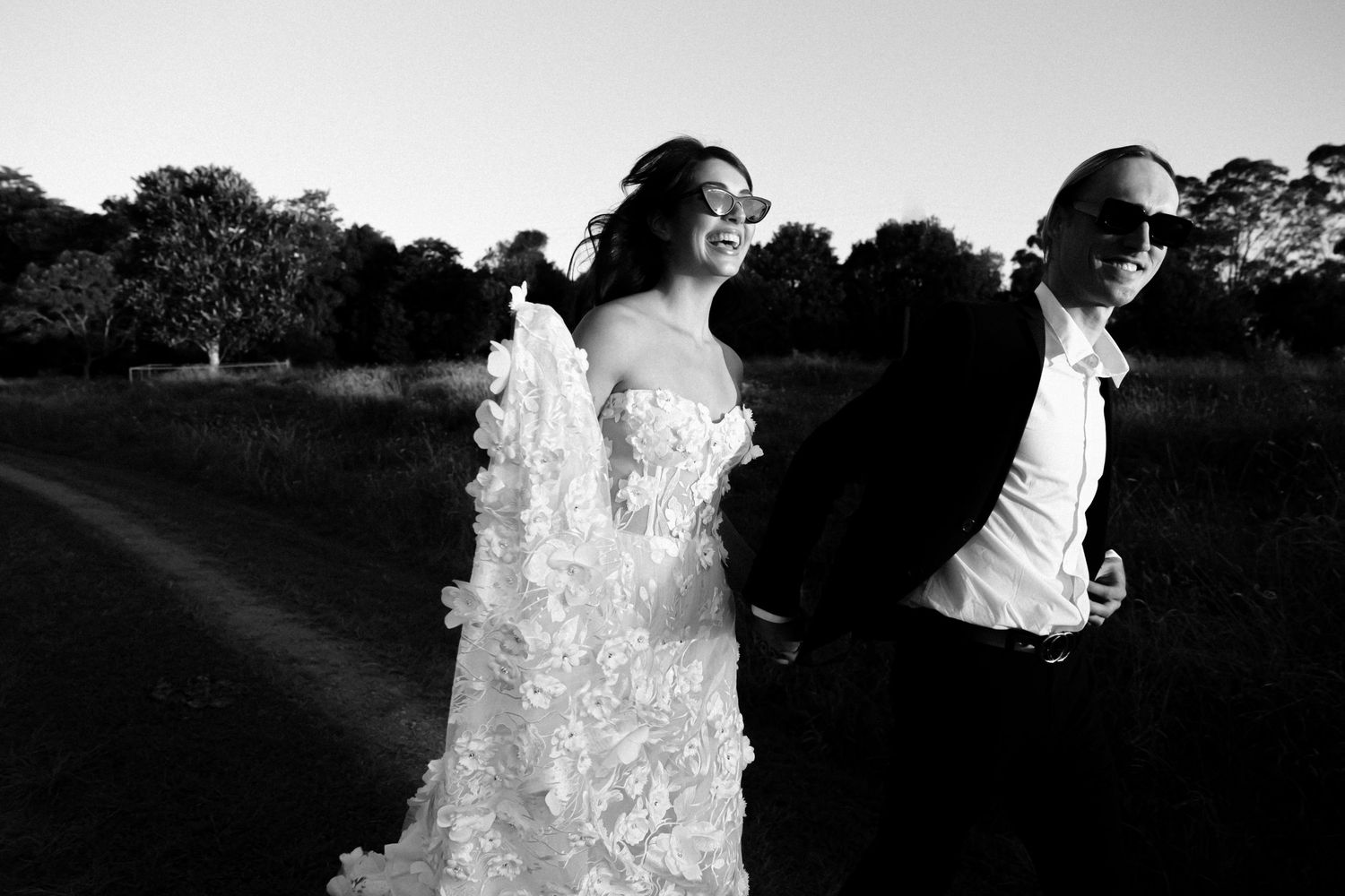 Black and white photo of a bride in a floral gown and groom in a suit walking outdoors at dusk.