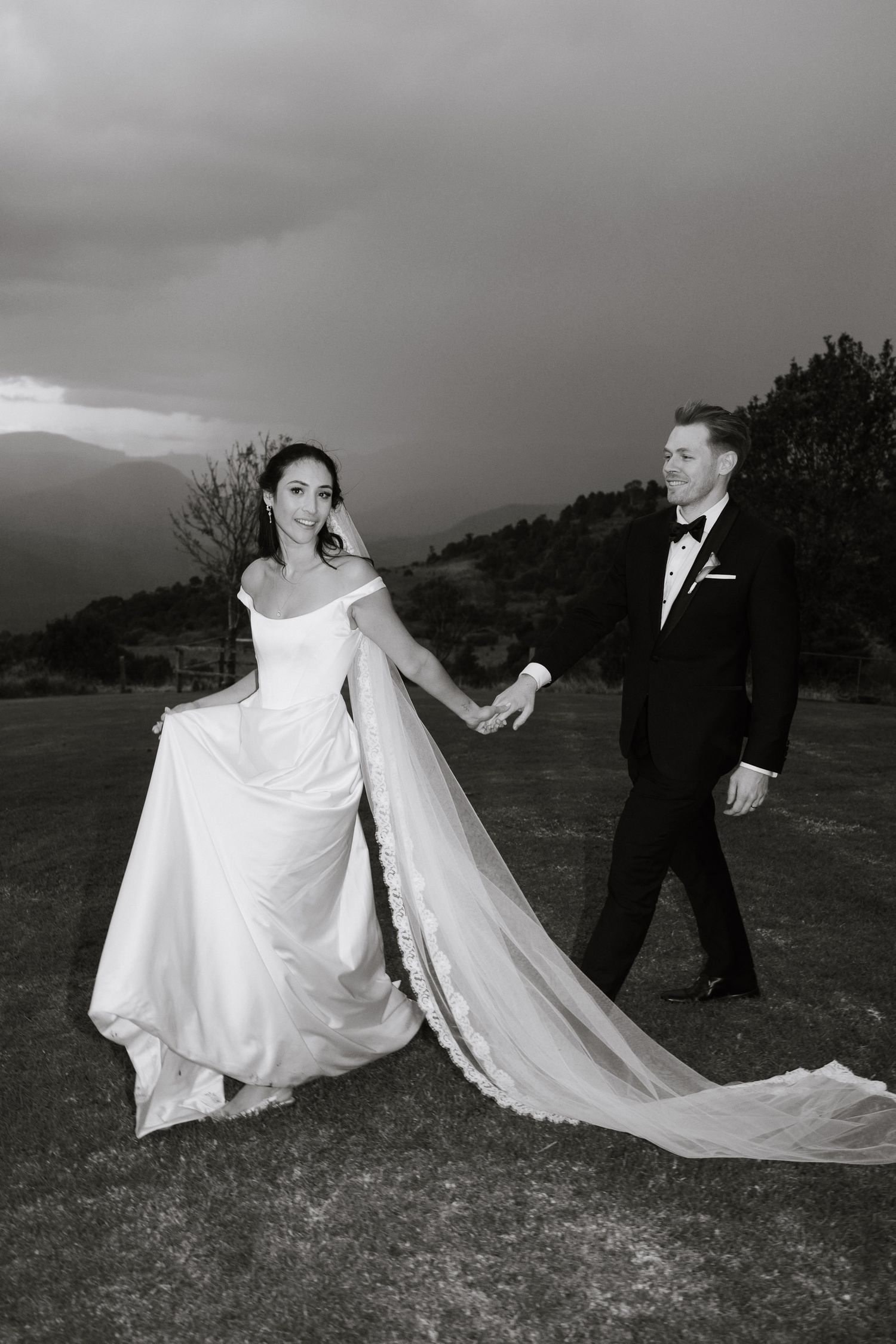 A couple in wedding attire playfully pose with a flowing white veil against a scenic hillside backdrop.