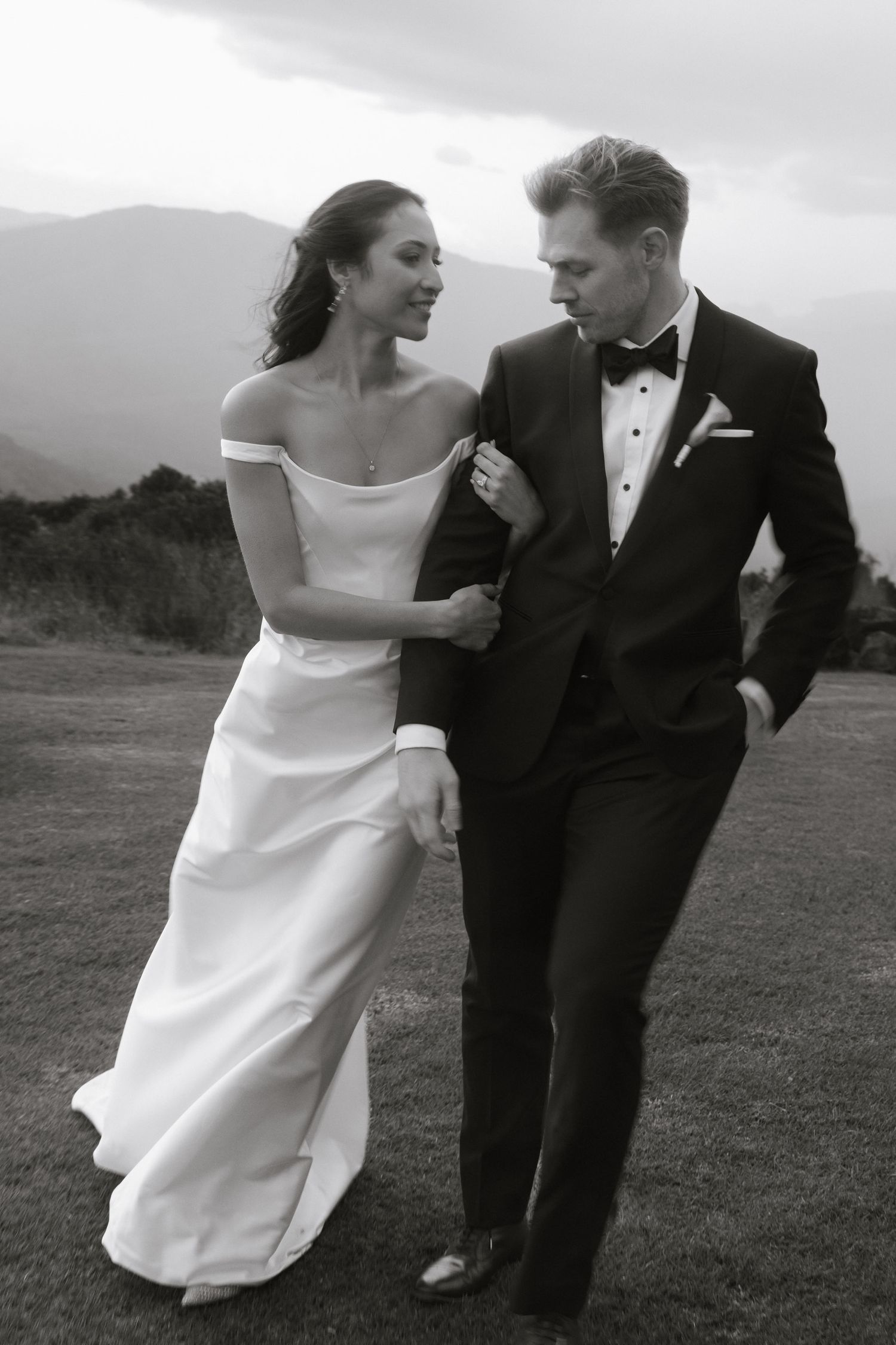 Classic black and white wedding photo of couple walking together outdoors with mountain scenery in the background.