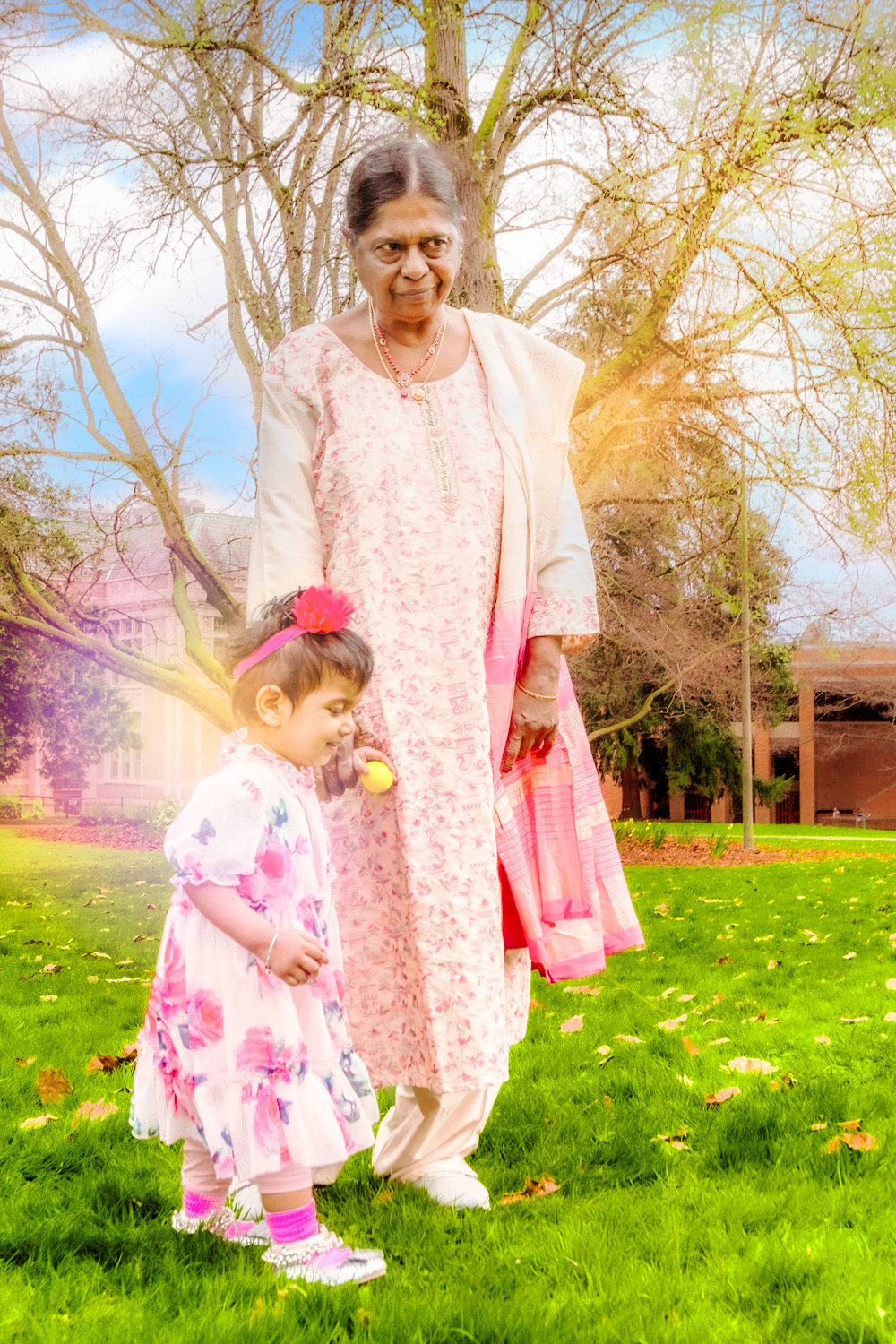 A grandmother and small child in matching pink dresses stand together on a sunny spring day in a grassy park.
