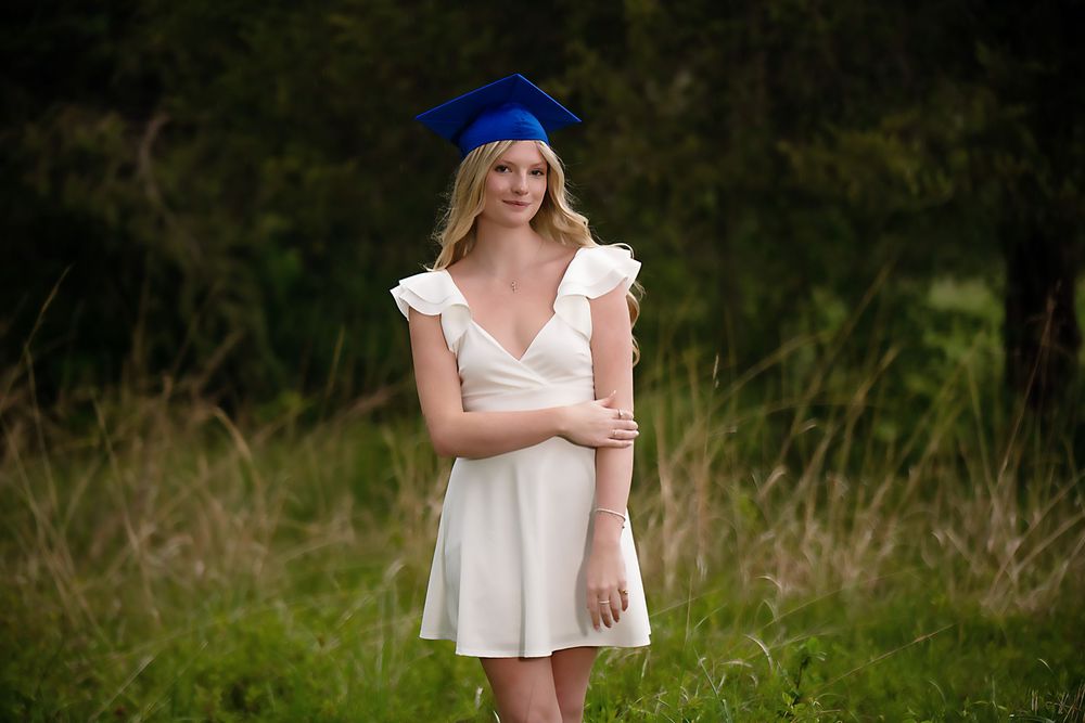 Graduate in white dress and blue cap poses in outdoor natural setting.