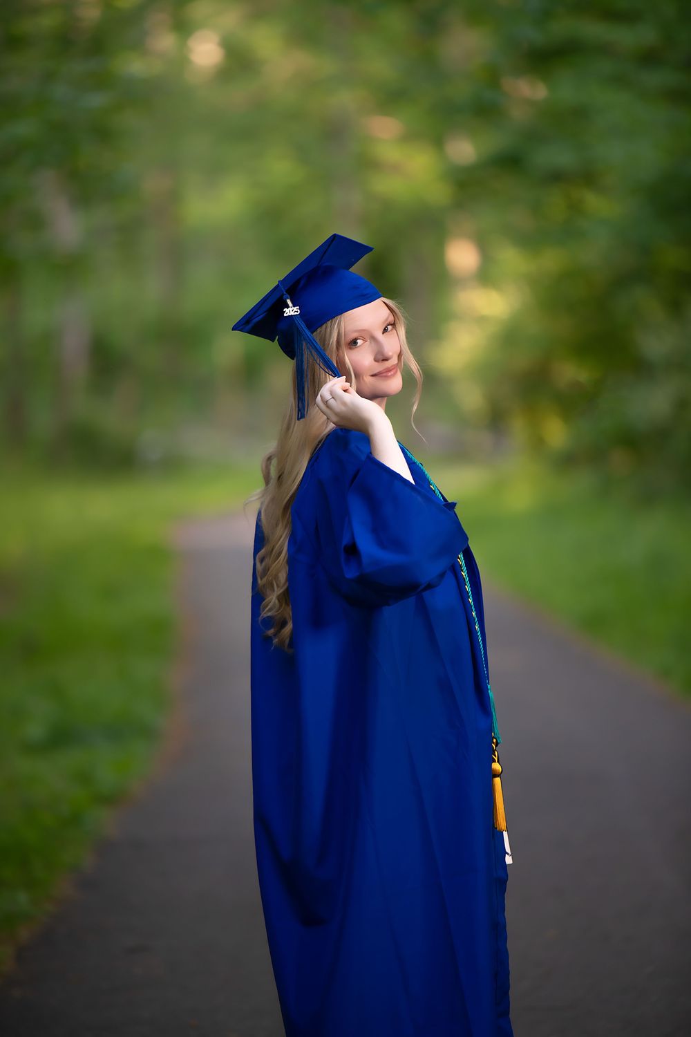 Graduate in blue cap and gown poses on pathway surrounded by green trees.