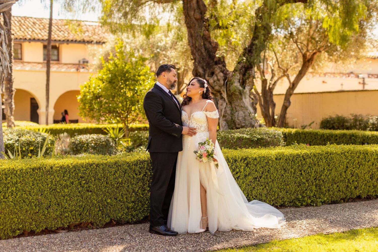 Couple sharing romantic moment in garden with manicured hedges and Mediterranean architecture.