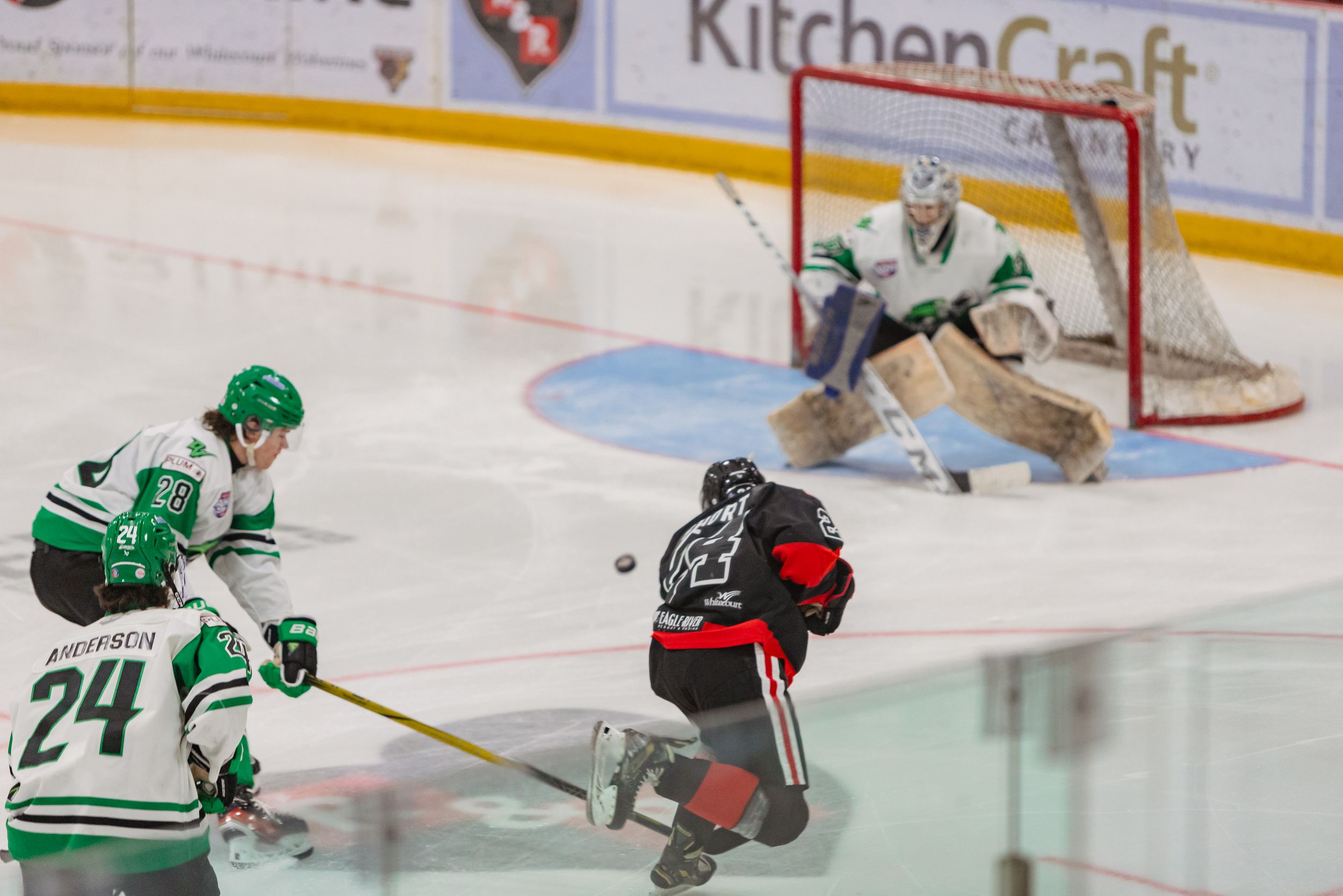 A hockey photo of a player in red and black shooting a puck at a goalie in white and green in front of the net. Two green/white players.