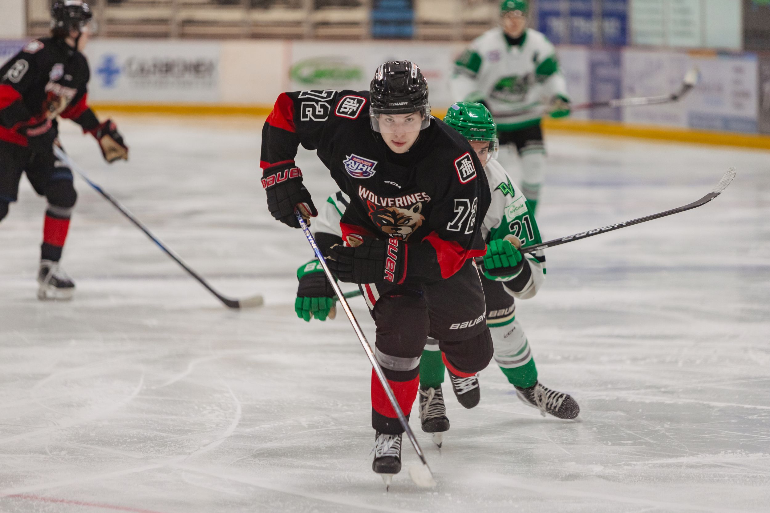 A hockey player in black and red skates towards the camera with a green and white player behind.