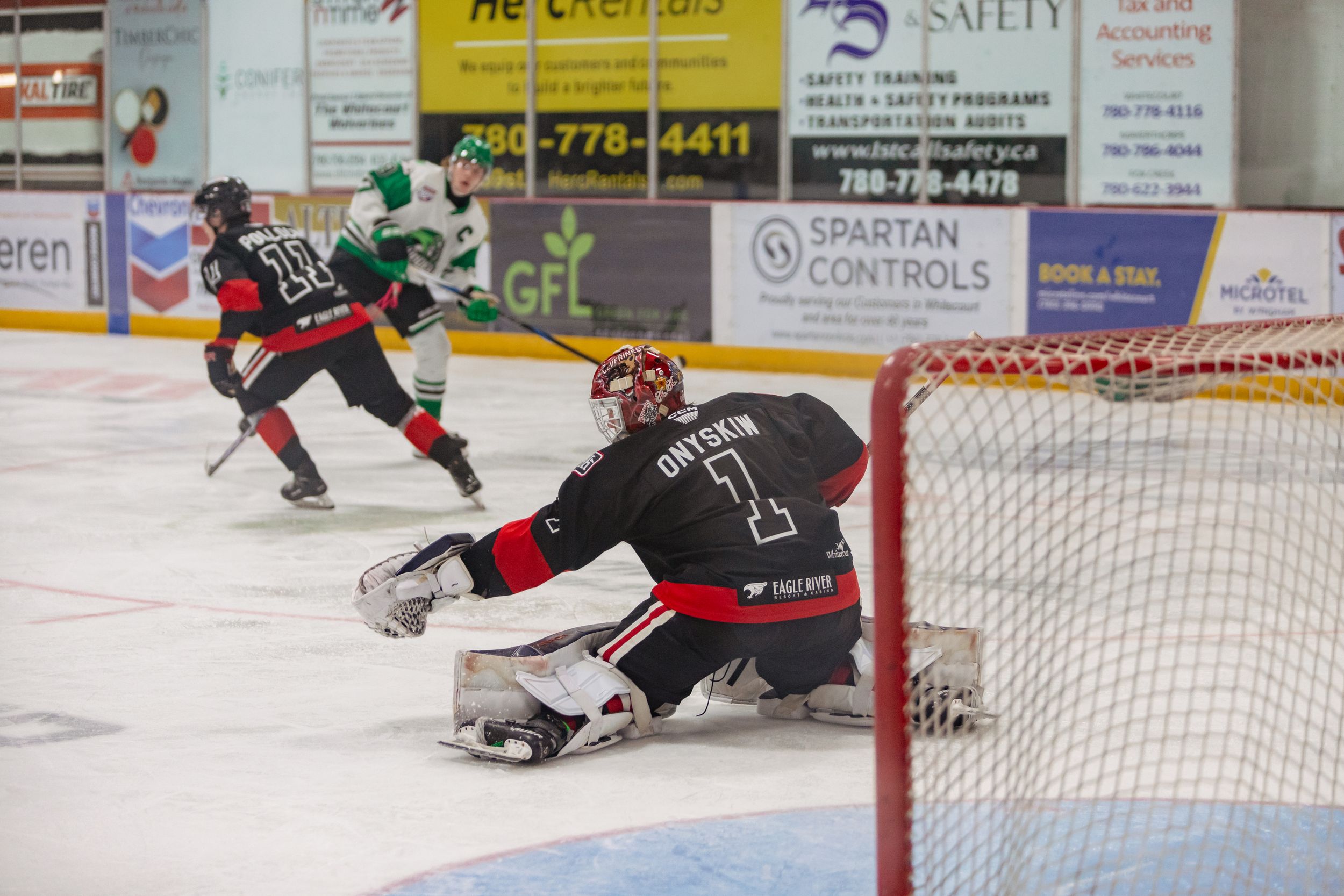 A hockey goalie in red and black catches a puck in front of the net. The player in green and white shoots it in the background.